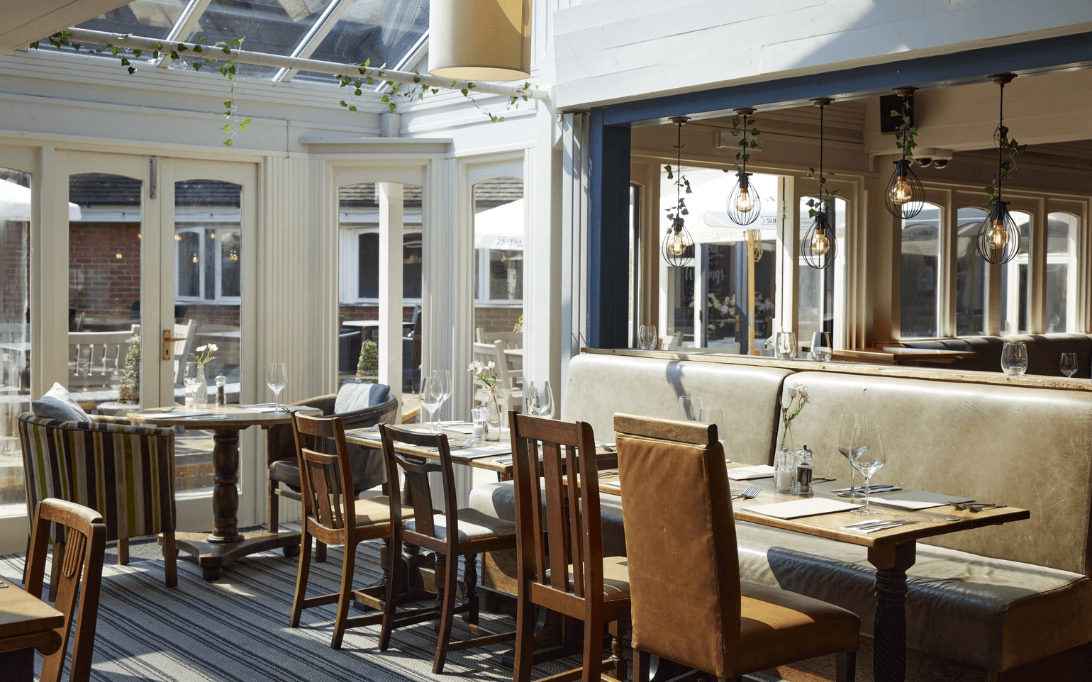 Sunlit conservatory dining area with wooden chairs, banquette seating and trailing ivy overhead.