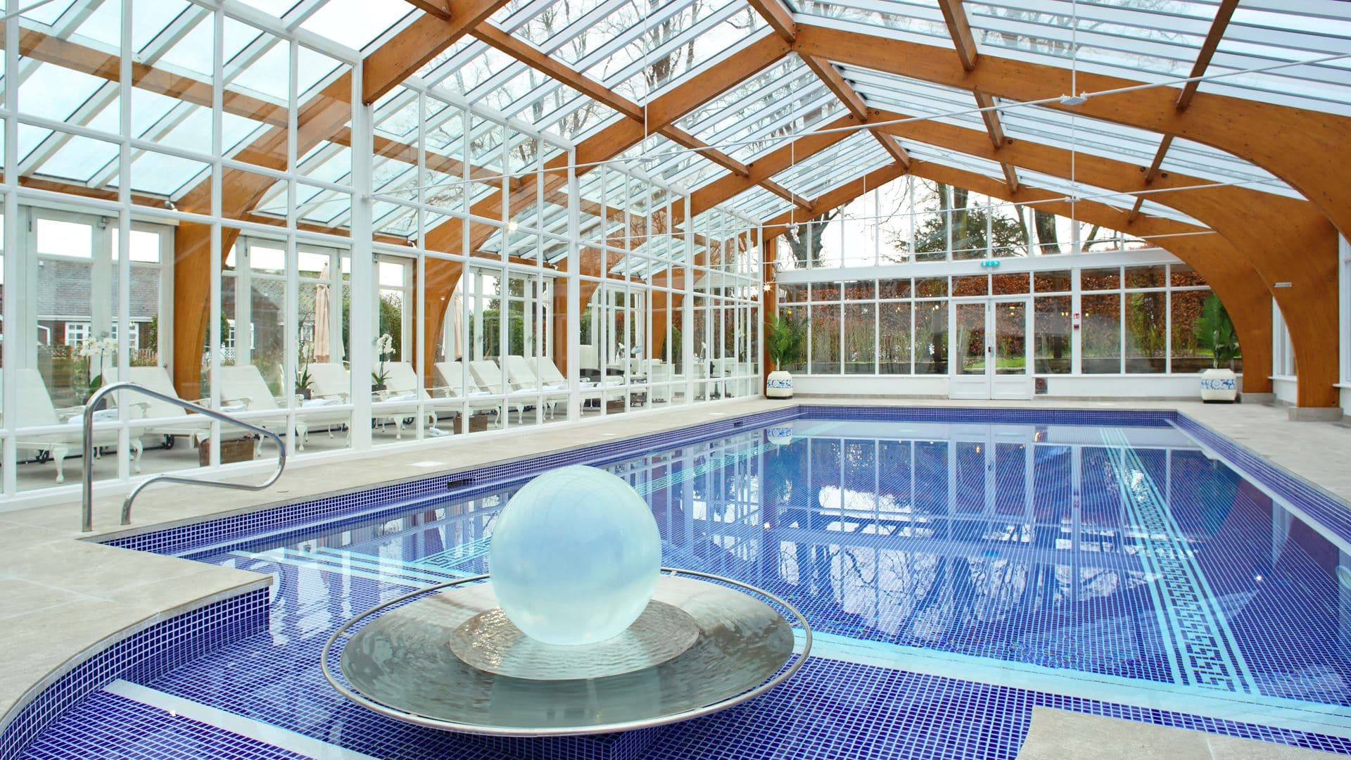 Indoor swimming pool with blue mosaic tiles beneath a glass and timber vaulted roof.