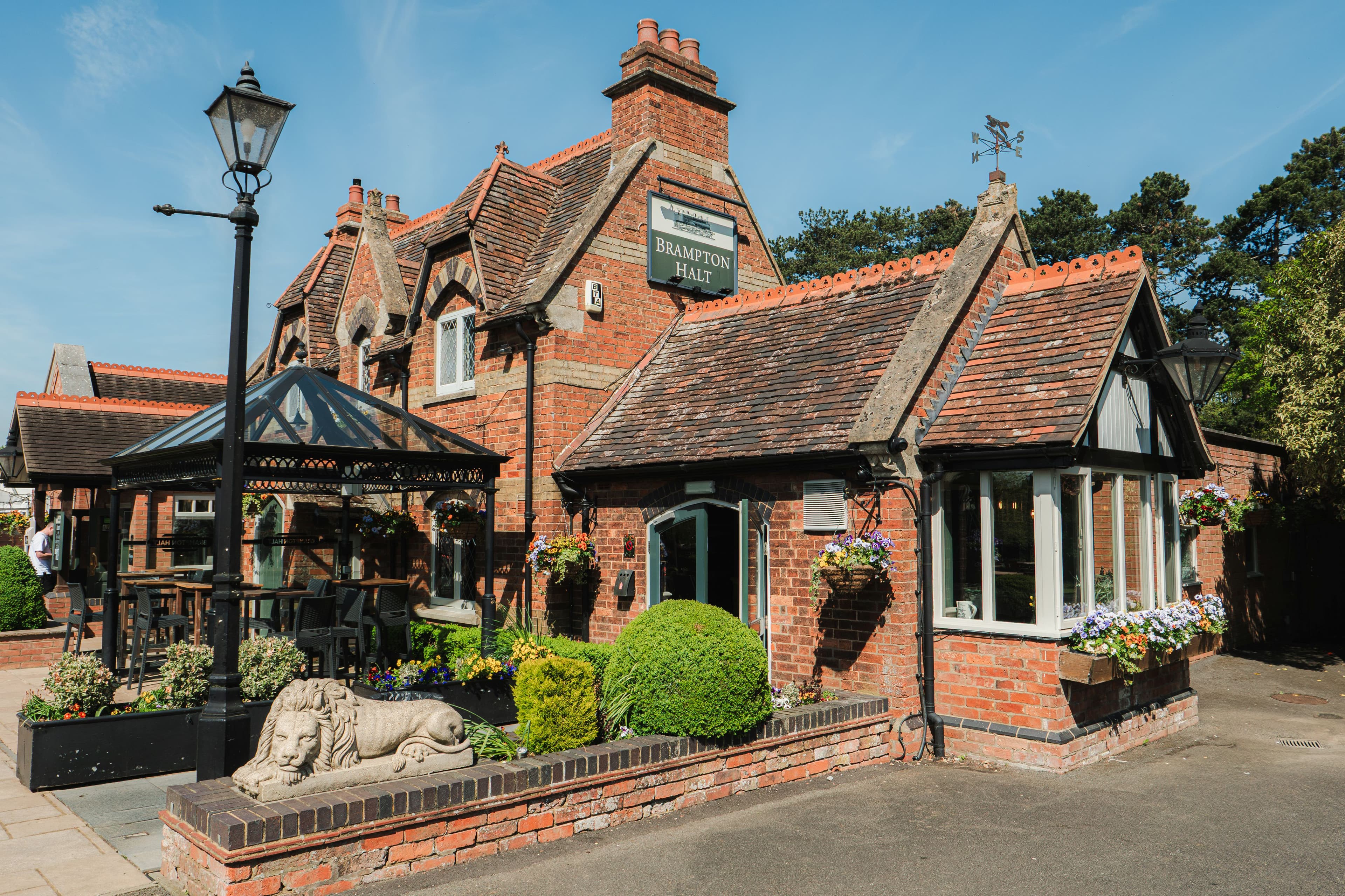 The red brick exterior of The Brampton Halt pub with hanging baskets and a stone lion.
