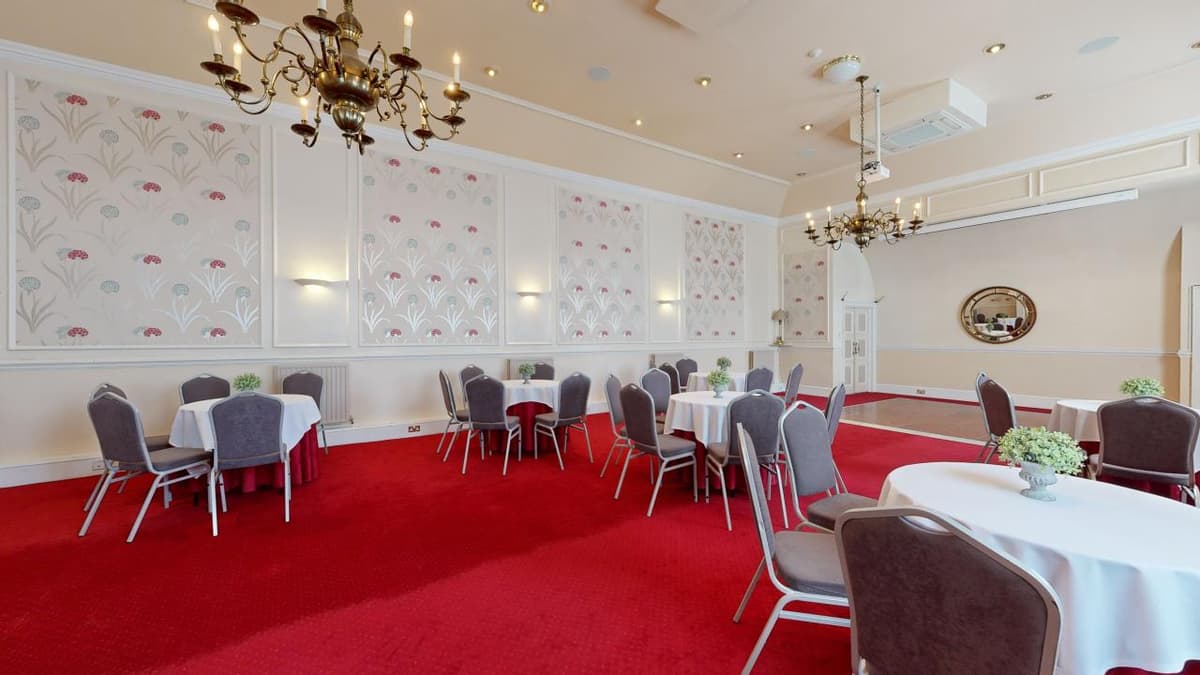 Function room with red carpet, round white-clothed tables, grey chairs and brass chandeliers.