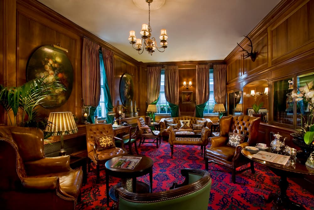 Wood-panelled lounge with tan leather armchairs, a chandelier and a red patterned carpet.