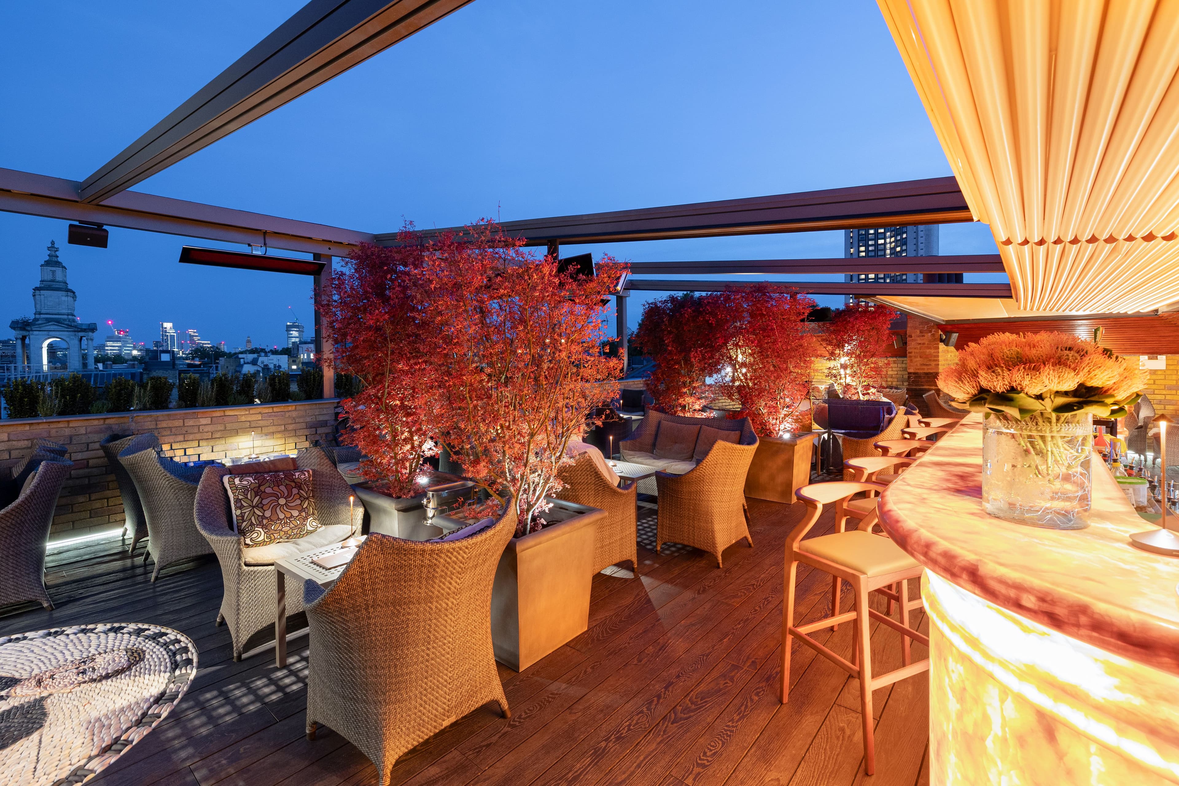 Rooftop terrace at dusk with wicker seating, red trees and London skyline views.