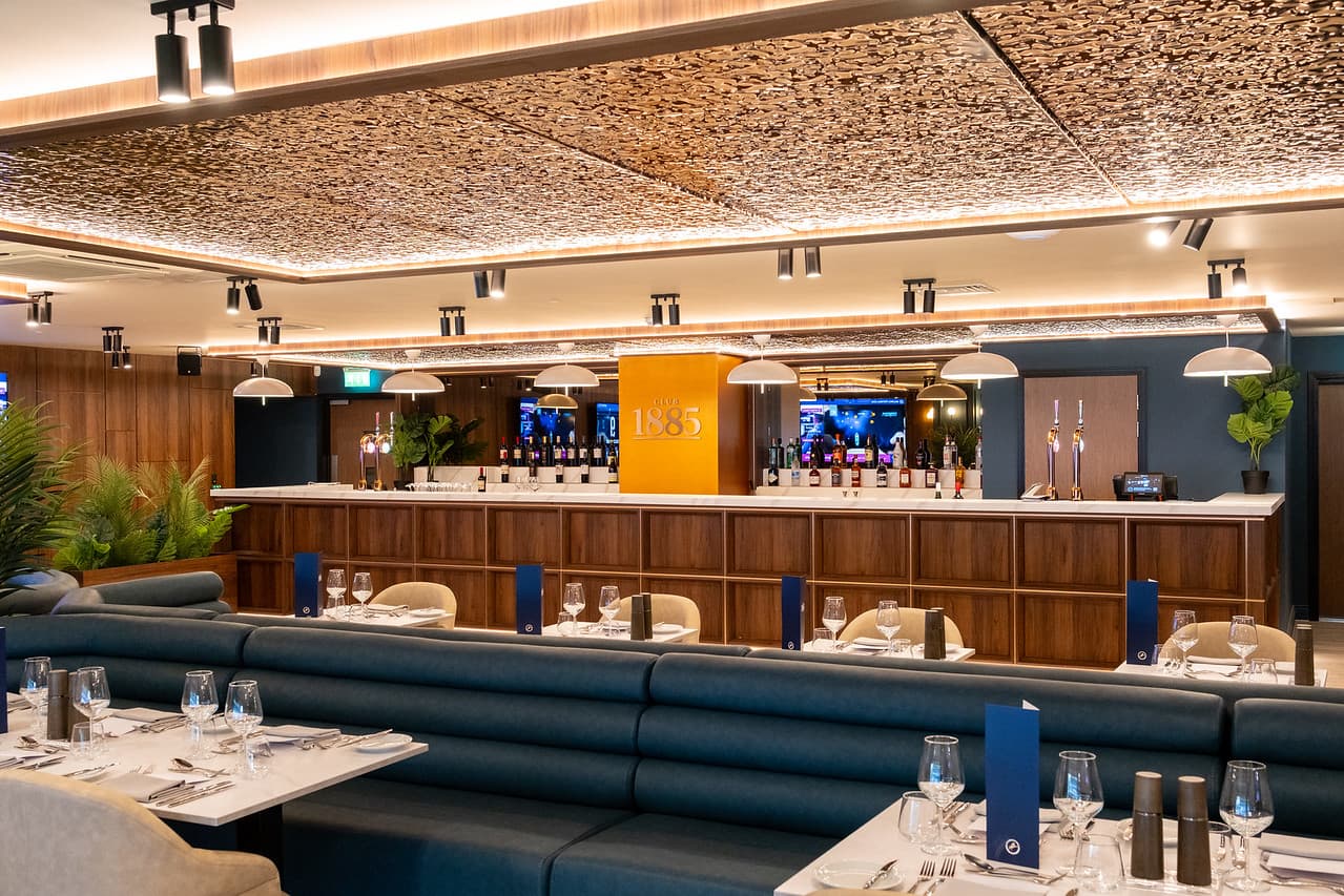 Club 1885 dining room with blue booth seating, laid tables and a wood-panelled bar.
