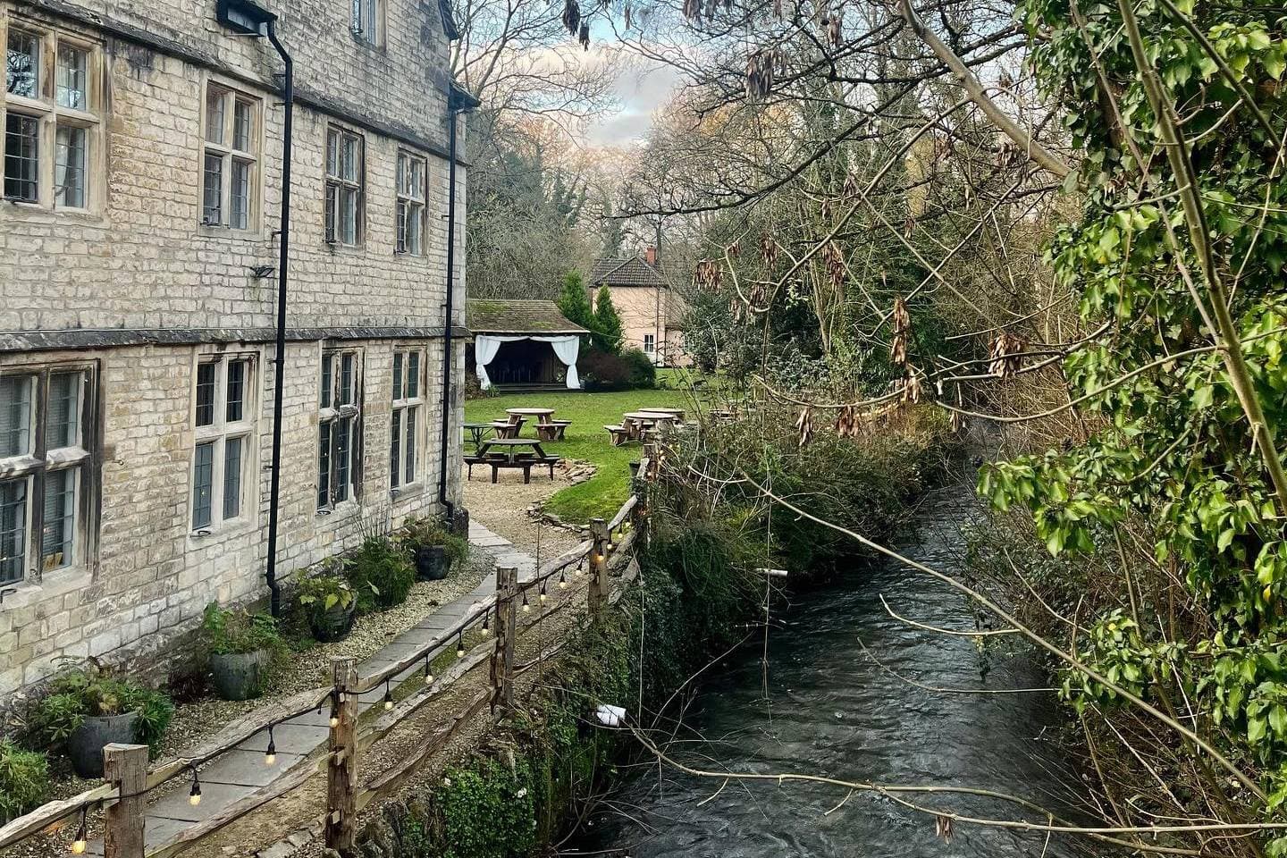 Stone mill exterior with riverside walkway, festoon lights and garden picnic tables.