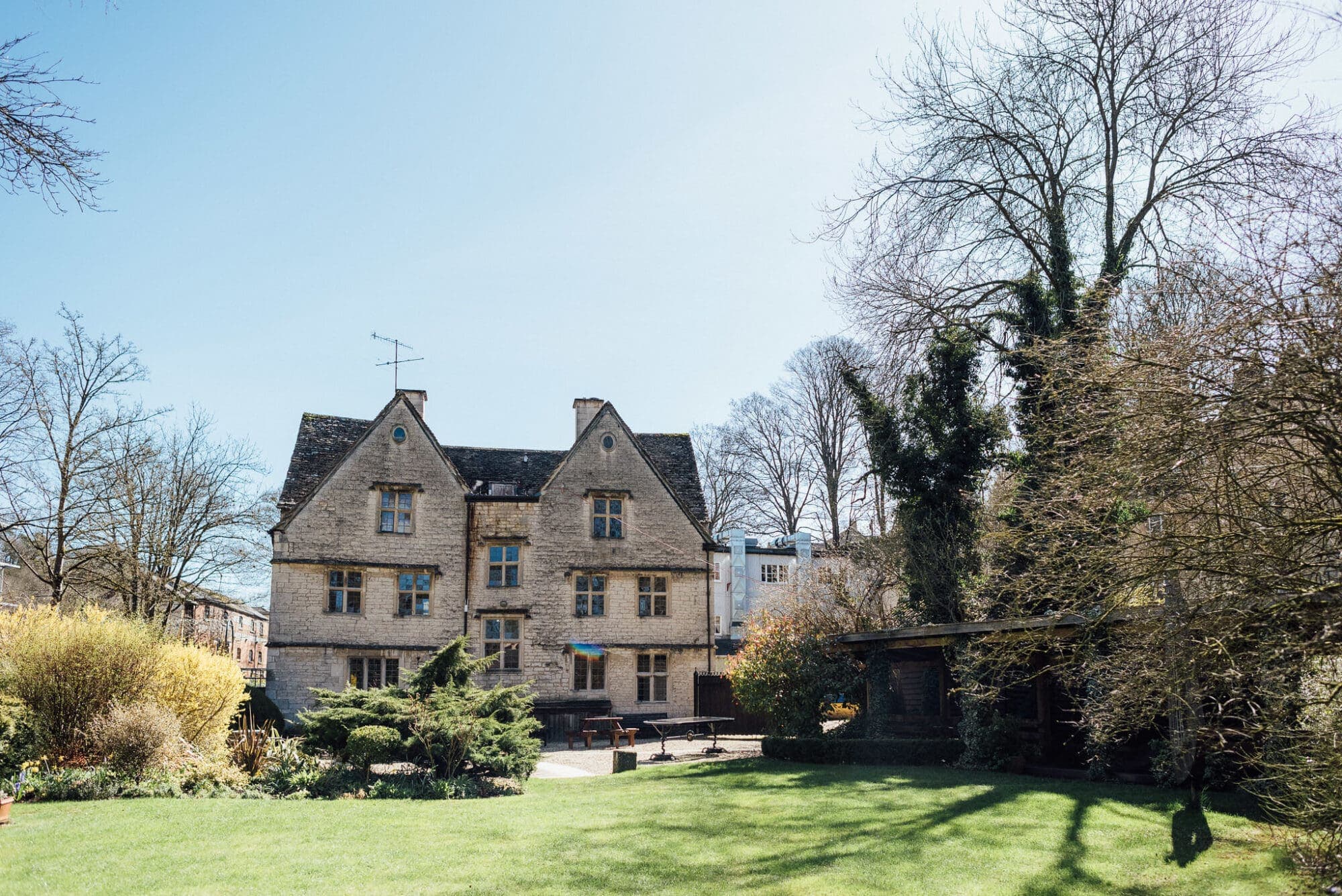Rear exterior of a Cotswold stone manor house with a lawned garden and bare trees.