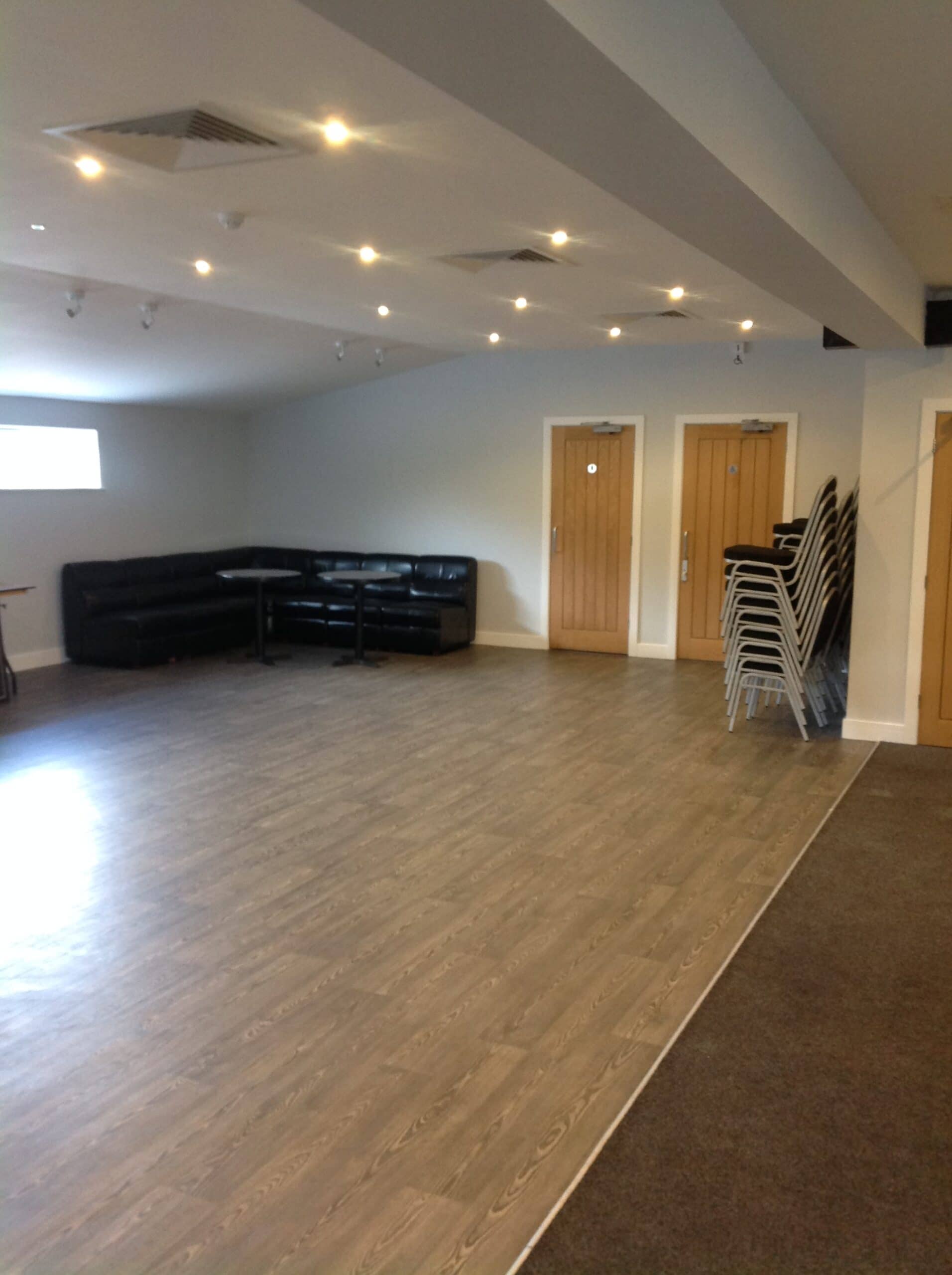 Spacious function room with wood-effect flooring, black leather seating and stacked chairs.