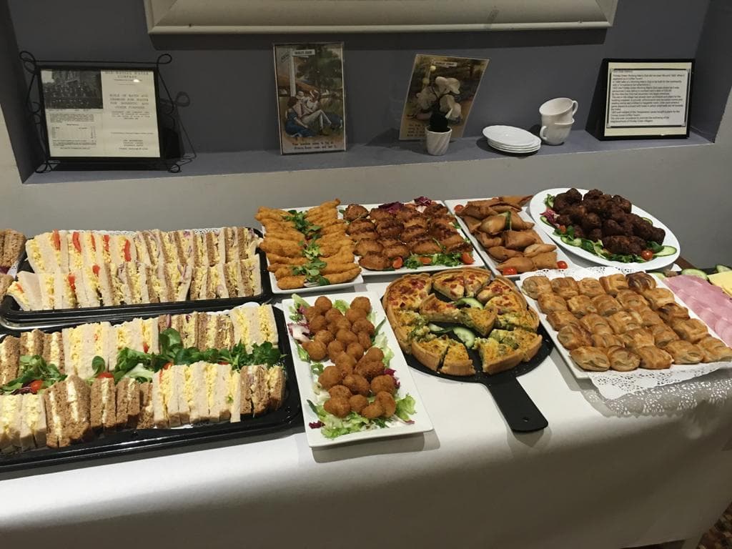 Buffet spread of sandwiches, quiche, sausage rolls and finger foods on a white-clothed table.