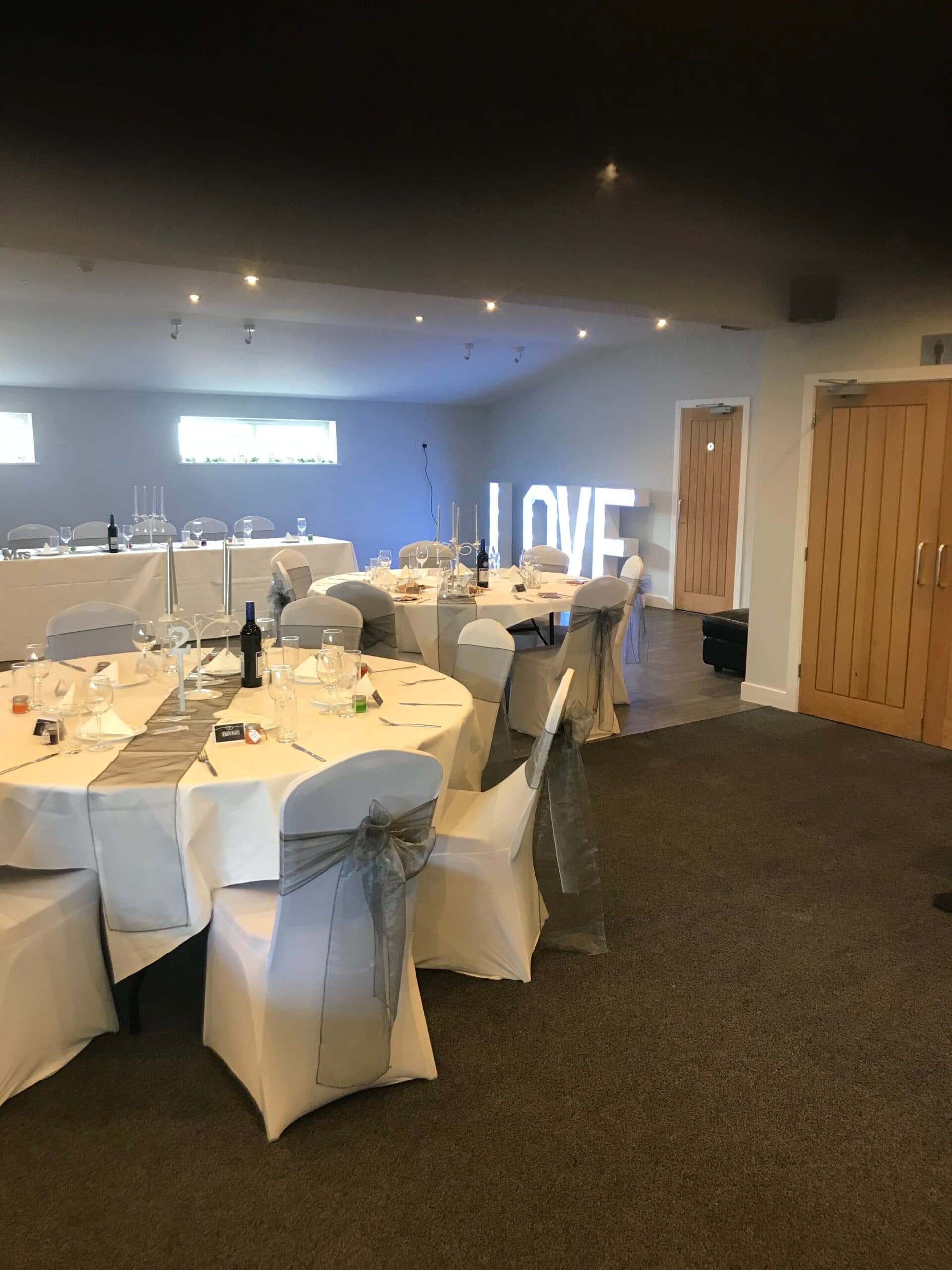 Function room laid for a wedding breakfast with white chair covers and a illuminated LOVE sign.