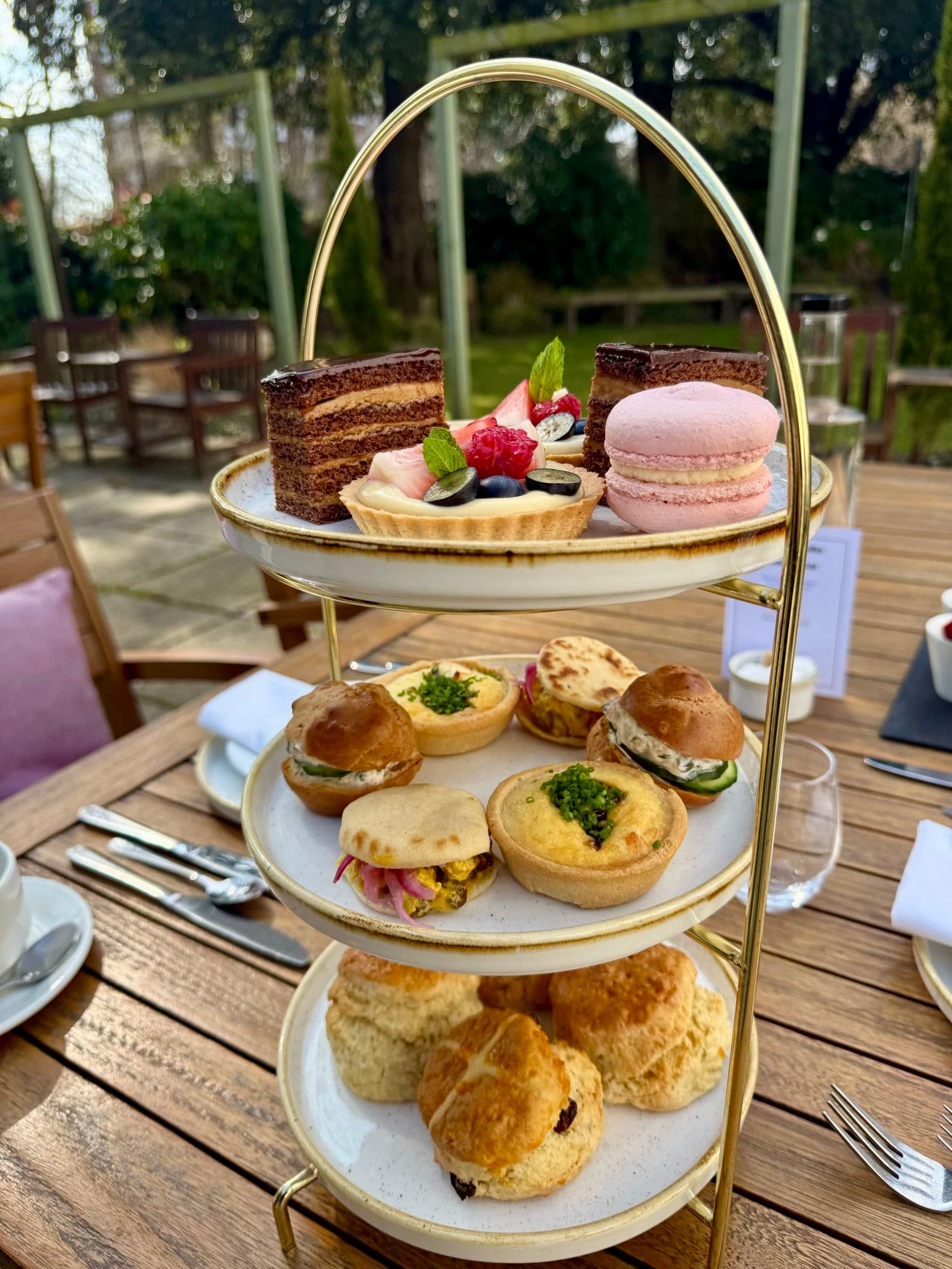 Three-tier gold afternoon tea stand with scones, savoury tartlets and cakes outdoors.