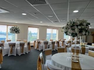 Function room laid for an event with white chair covers, gold sashes and tall floral centrepieces.