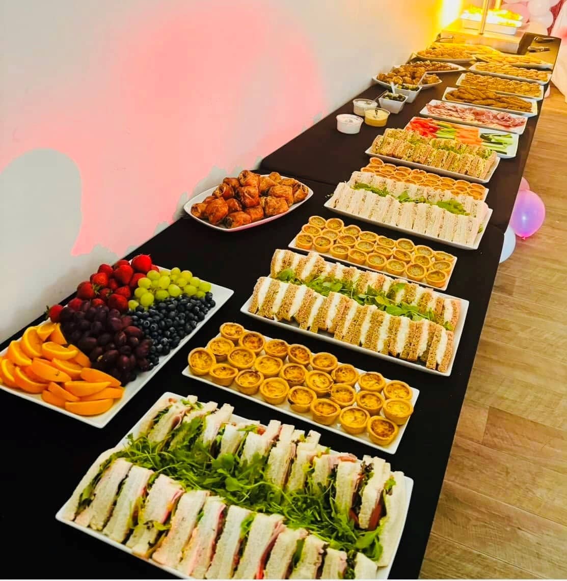 Long buffet table laid with sandwiches, mini quiches, fruit platters and party food.