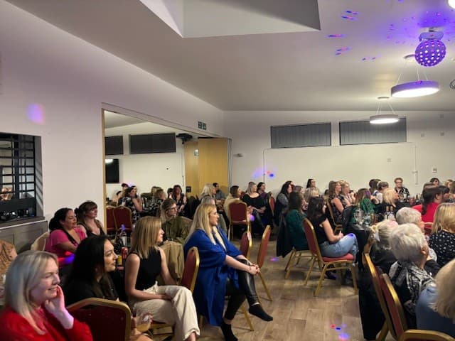 Rows of women seated in a function room lit with purple lighting.