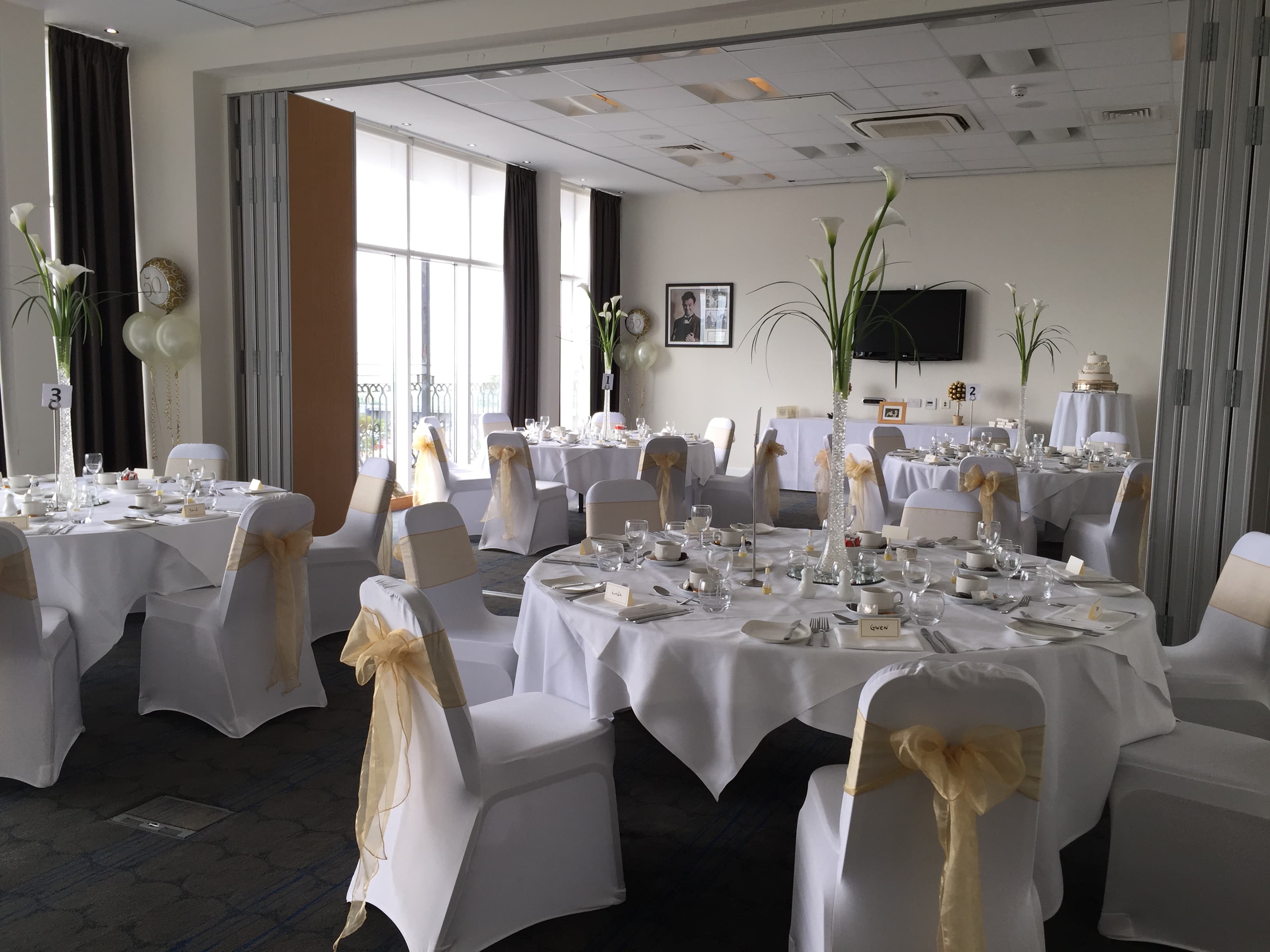 Function room laid for a celebration with white chair covers and gold sashes.
