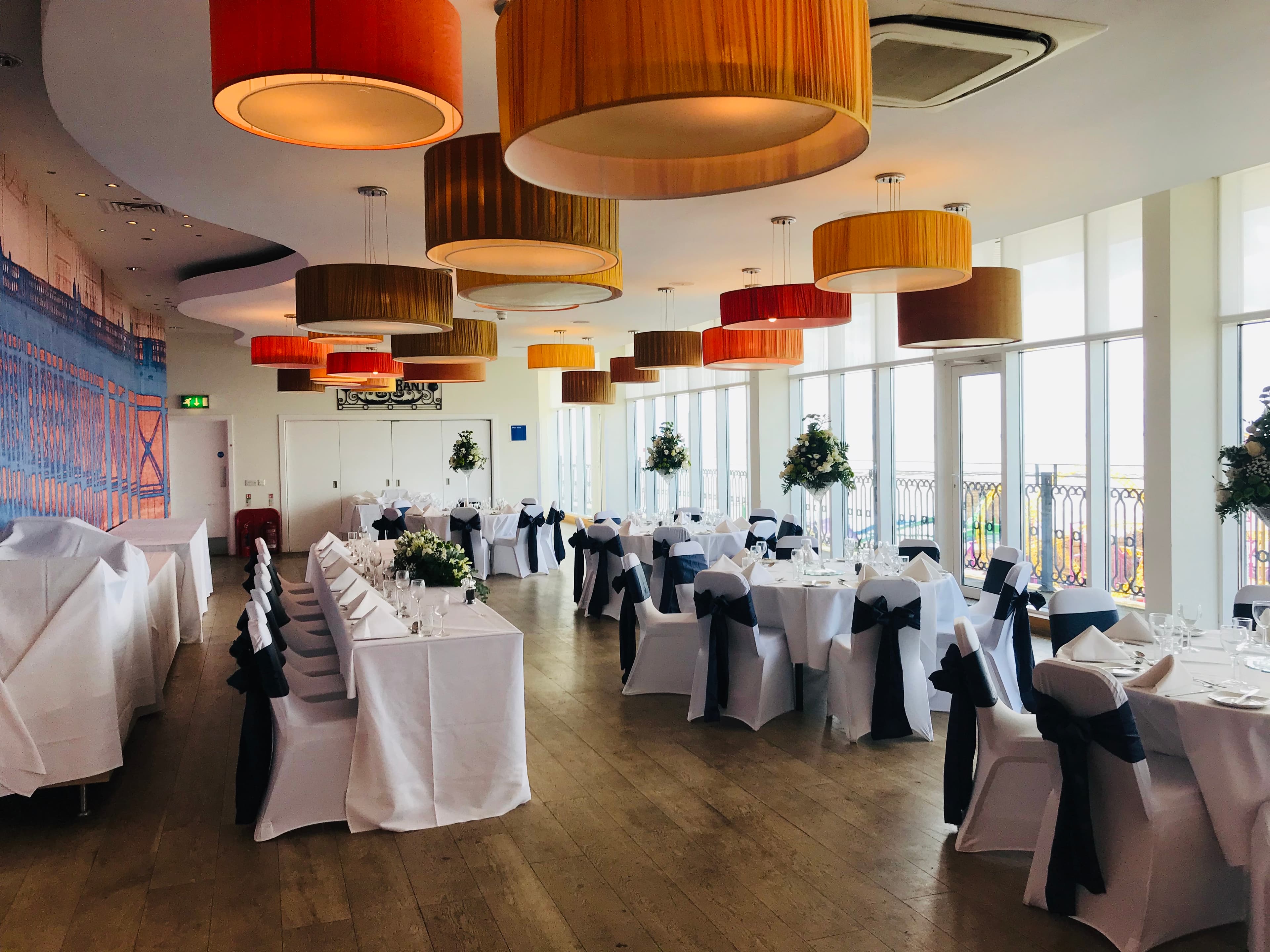 Bright function room with white-draped chairs, black sashes and tall floral centrepieces overlooking the seafront.