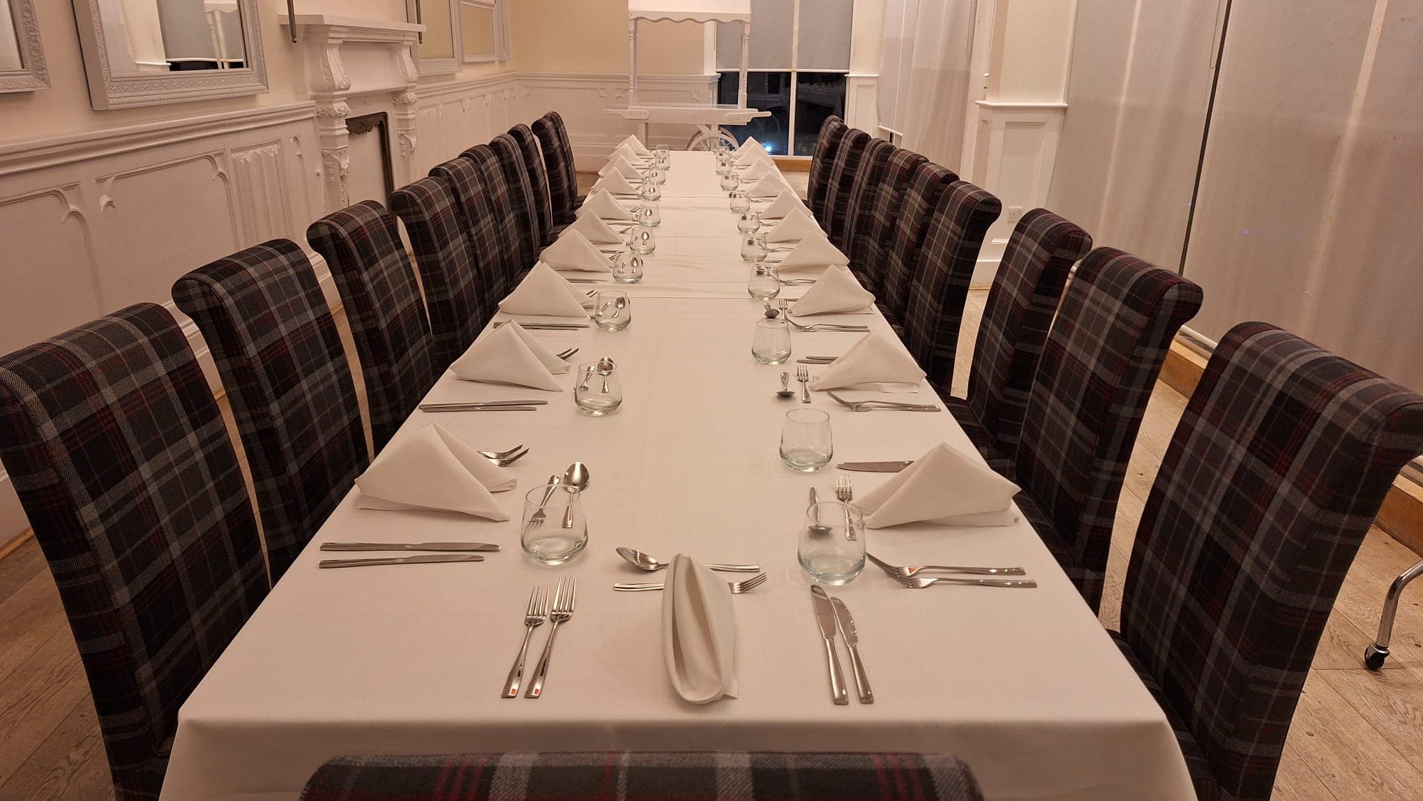 Long banquet table laid with white linen, tartan chairs and folded napkins in a private dining room.