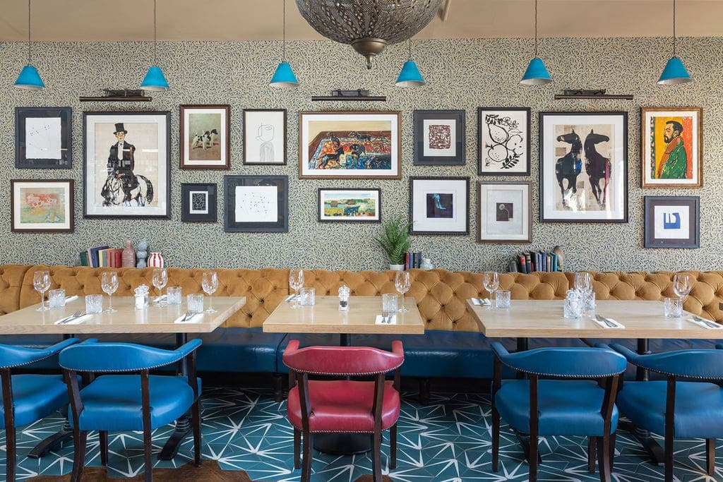 Restaurant dining area with tufted banquette seating, blue chairs and a gallery wall of framed artwork.