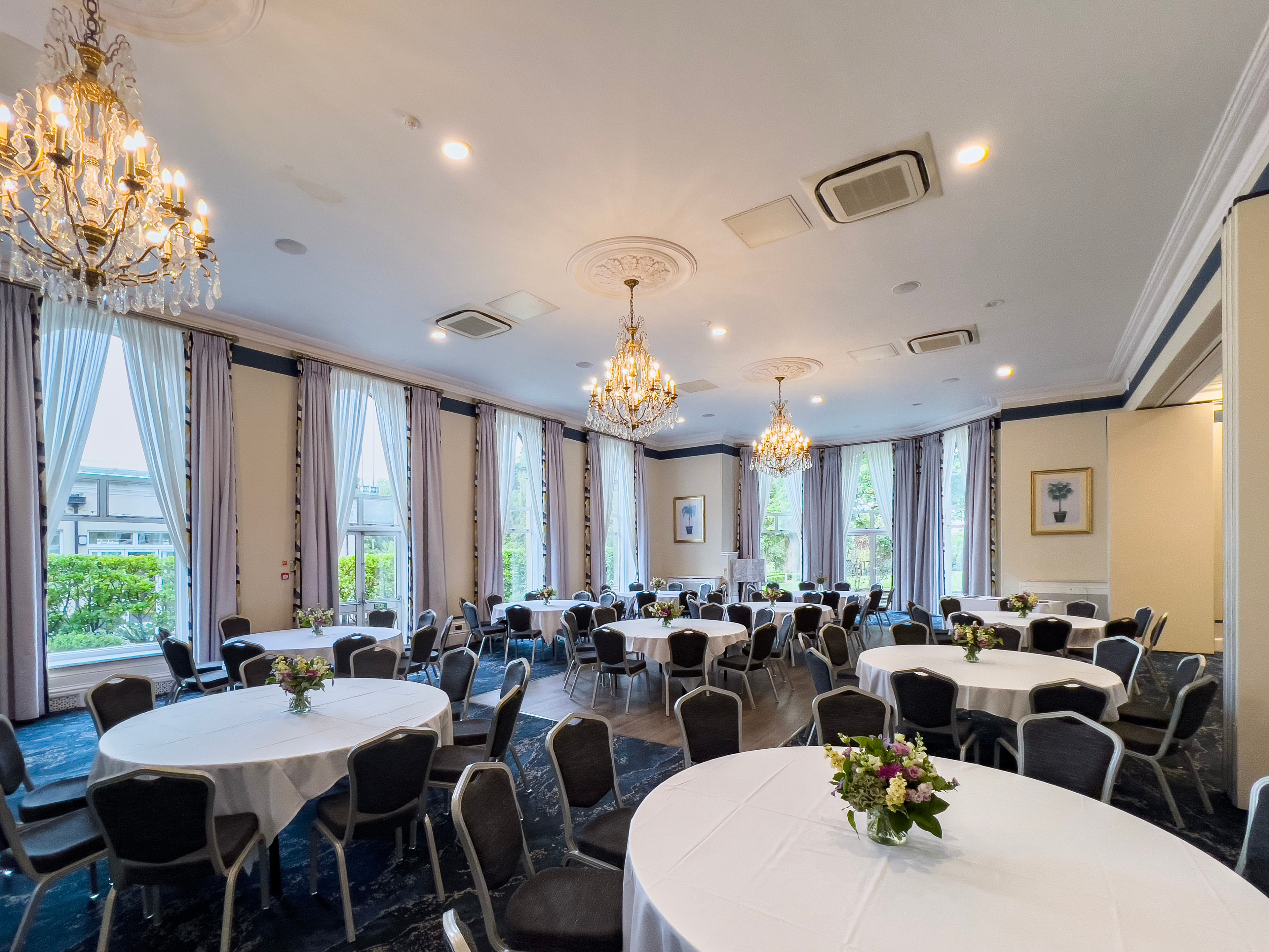 Bright function room with round white-clothed tables, chandeliers and tall garden-facing windows.