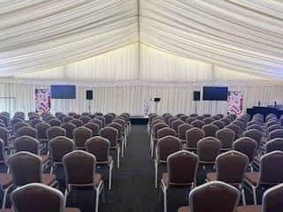 Rows of banqueting chairs inside a large white marquee with two presentation screens.