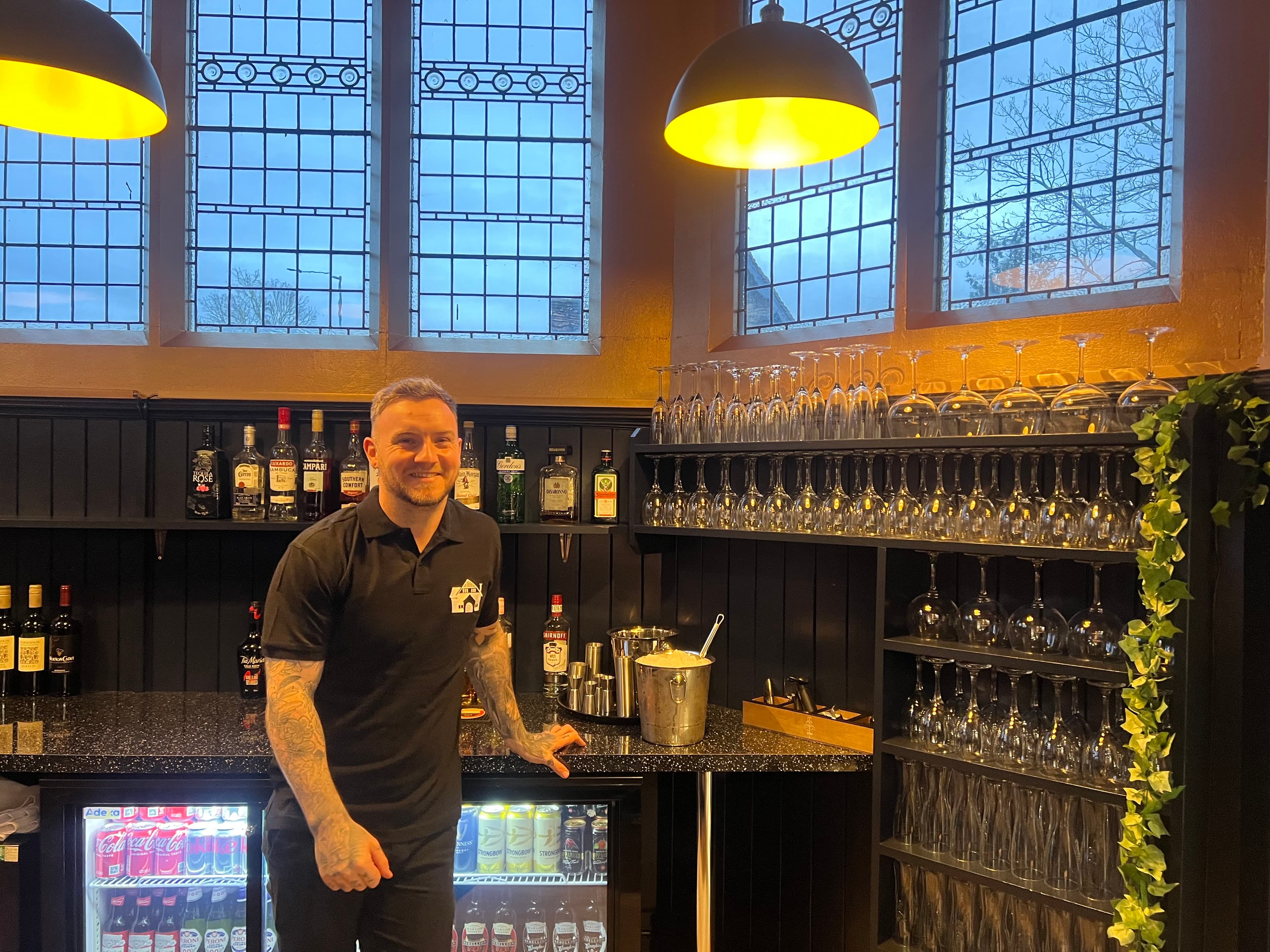 Bartender standing behind the bar at Waddesdon Hall, with stocked shelves and wine glasses.