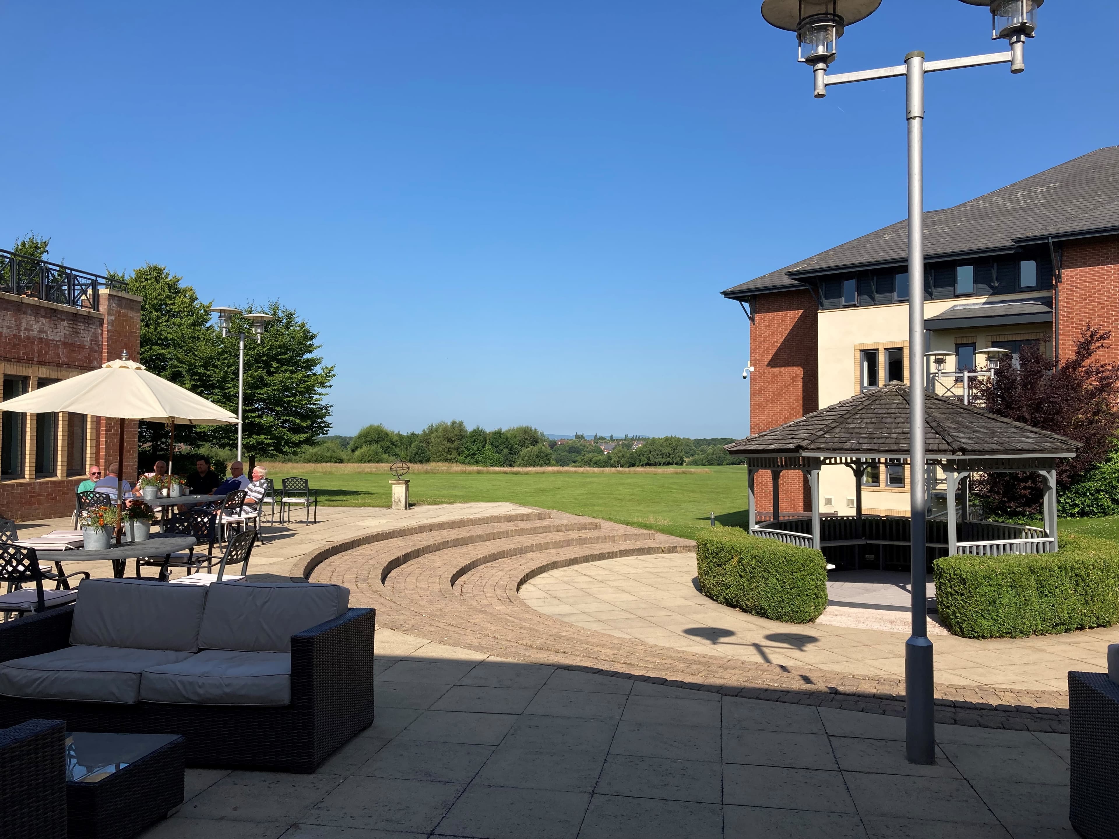 Sunny paved terrace with outdoor seating, a gazebo and open lawned grounds beyond.